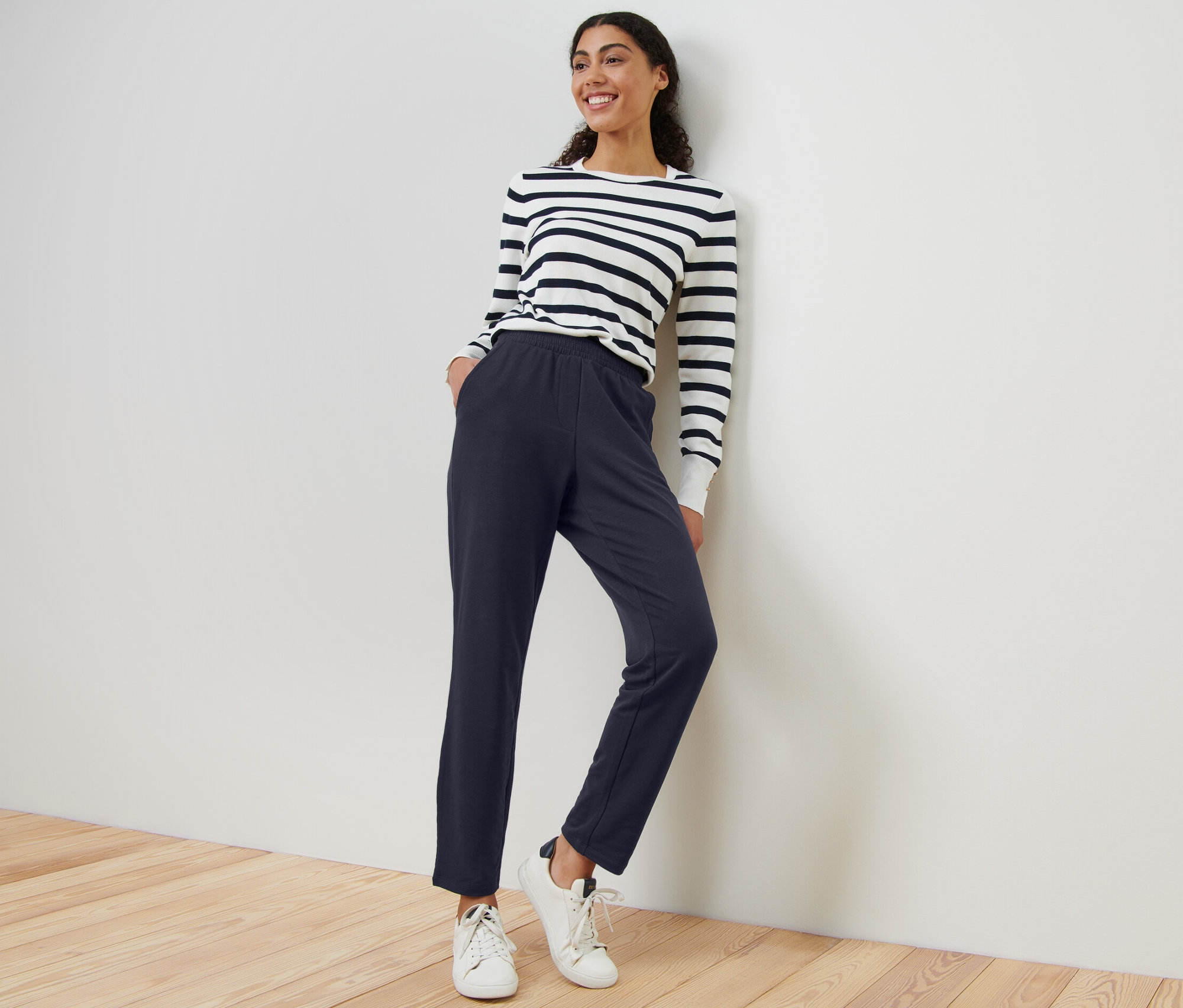 Nő sötétkék női lezser nadrágot és csíkos felsőt visel, miközben a falnak támaszkodik.