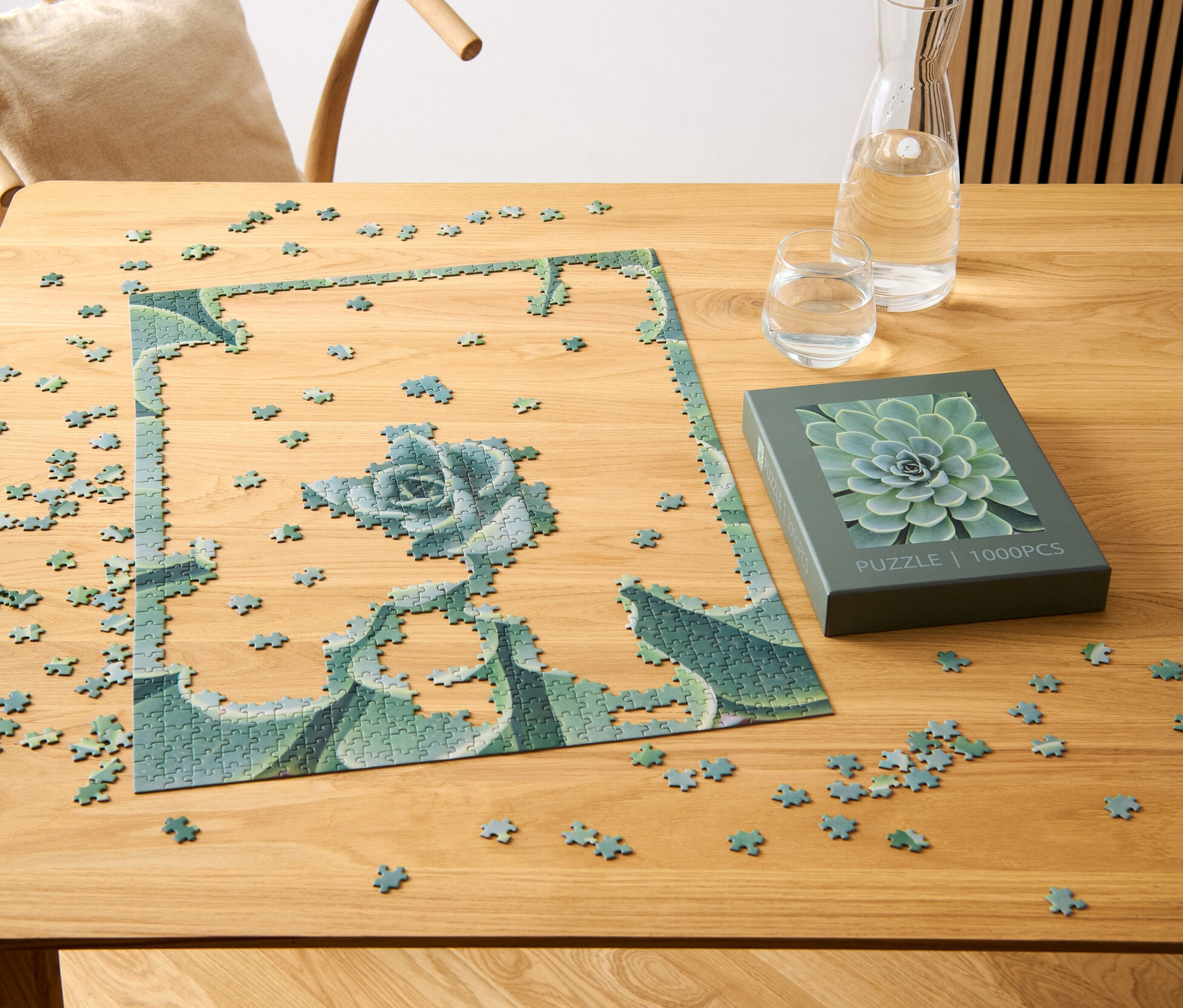 Egy részben összerakott, pozsgás növény mintájú puzzle egy fa asztalon van, egy pozsgás növény motívummal ellátott doboz, egy pohár és egy vízzel teli kancsó mellett.