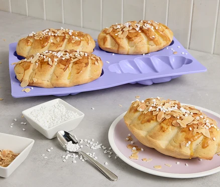 Négy mini fonott kalács egy lila sütőformában, egy tányéron, mandulával és cukorral megszórva.