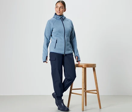 Egy nő egy fa szék mellett áll, kék kapucnis pulóvert és Női softshell lezser nadrágot visel.