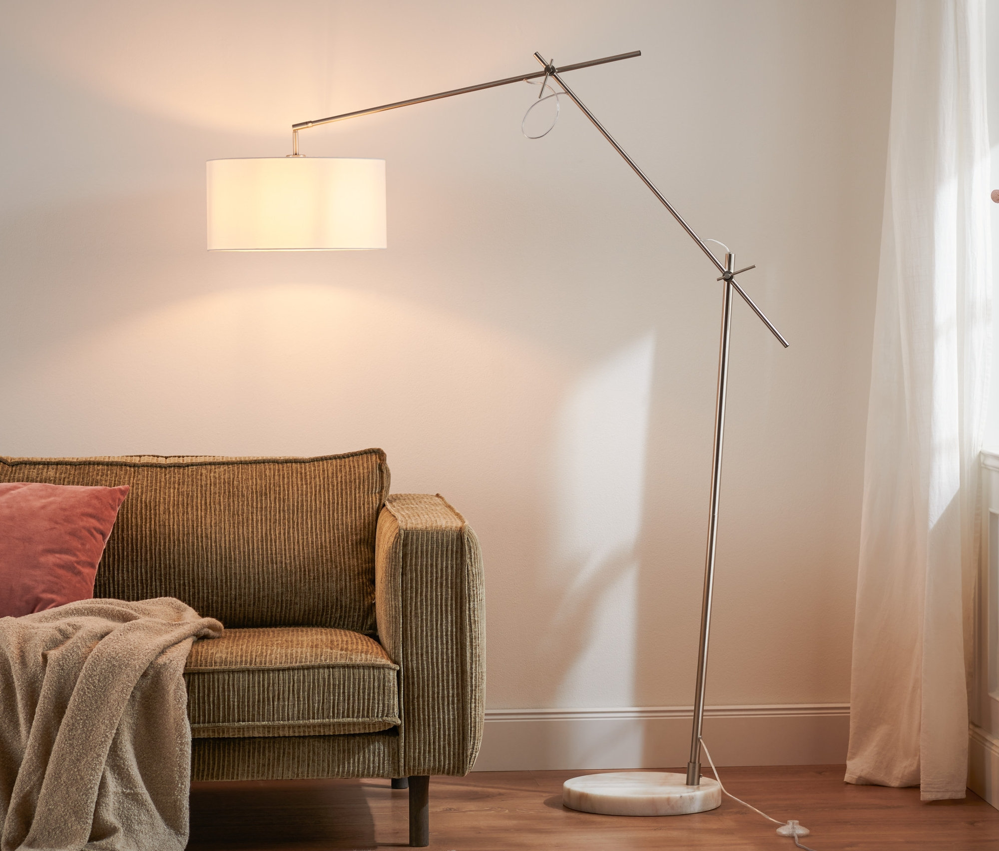 Egy barna kanapé rózsaszín párnával és bézs takaróval, mellette egy textilernyős állólámpa áll.