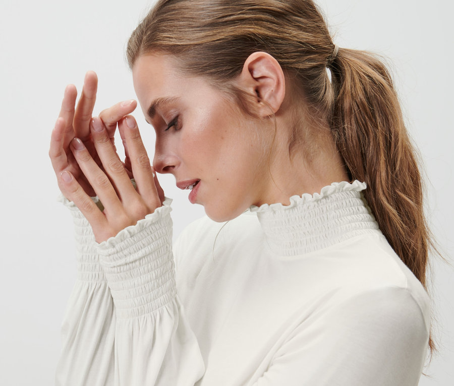 Egy nő csukott szemmel érinti meg az arcát. Krém színű, állógalléros ingblúzt visel.