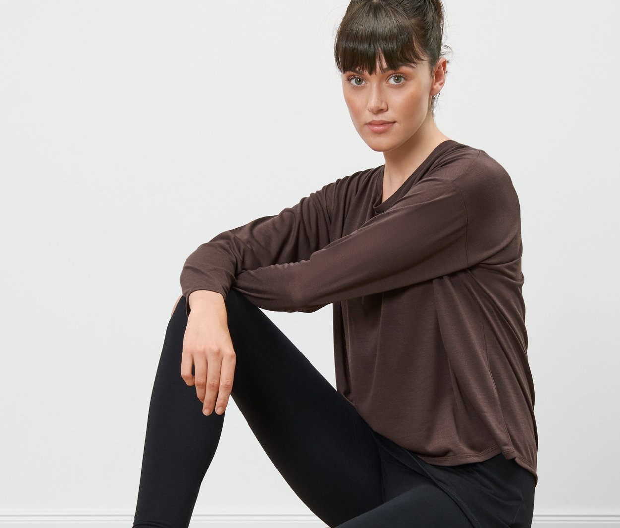 Nő ül egy fa ülőkén, barna/fekete 2 az 1-ben sportfelsőt és fekete leggingst visel.