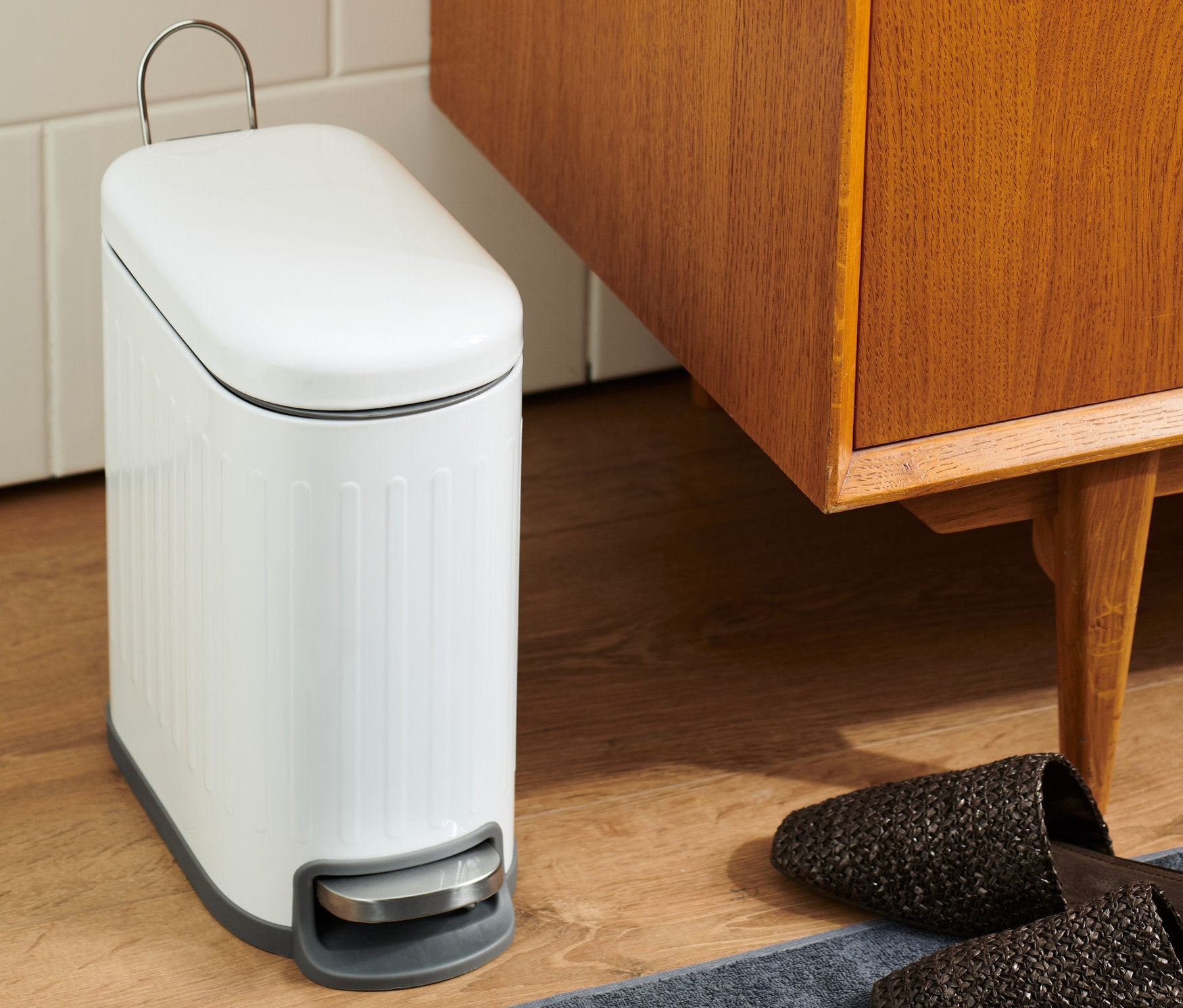 A fürdőszobában egy fehér fürdőszobai szemetes áll egy barna fa szekrény és papucsok mellett egy szürke szőnyegen.