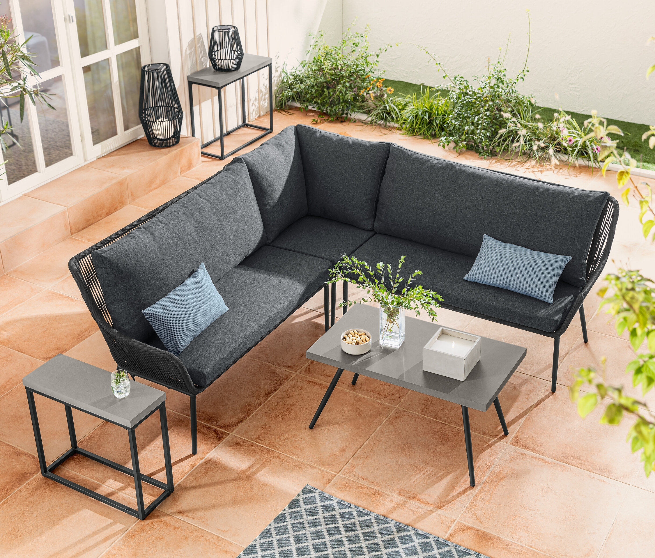 A textilfonatos lounge-sarokgarnitúra a policement asztallapos kisasztal mellett áll. A policement asztallapos konzolasztal-szett a textilpántos napelemes viharlámpa mellett található.