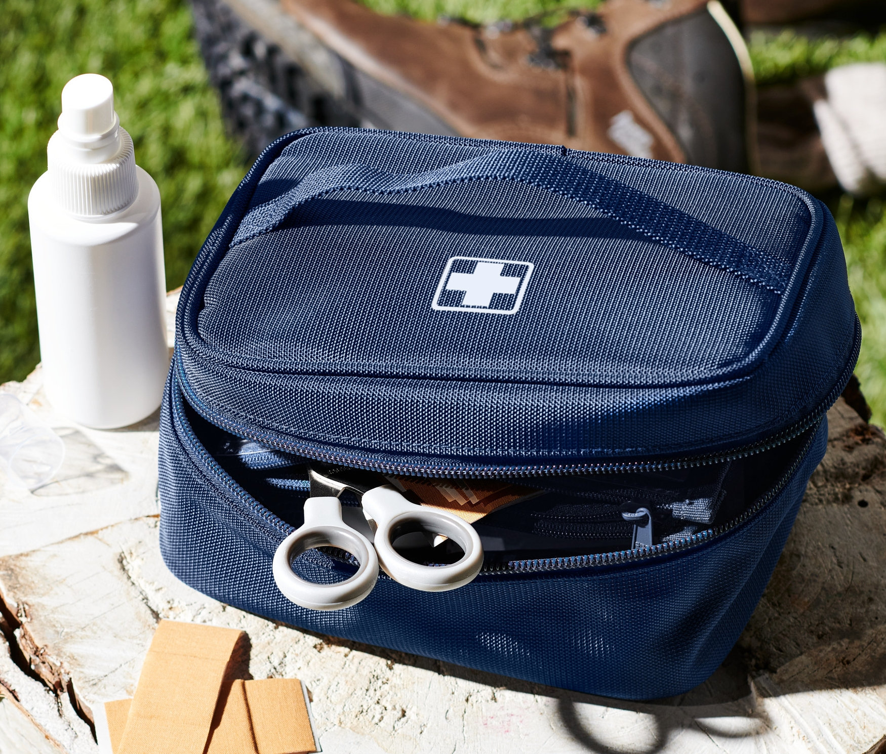 Kék gyógyszeres táska fehér kereszttel egy fatönkön, spray, tapaszok és cipők mellett.