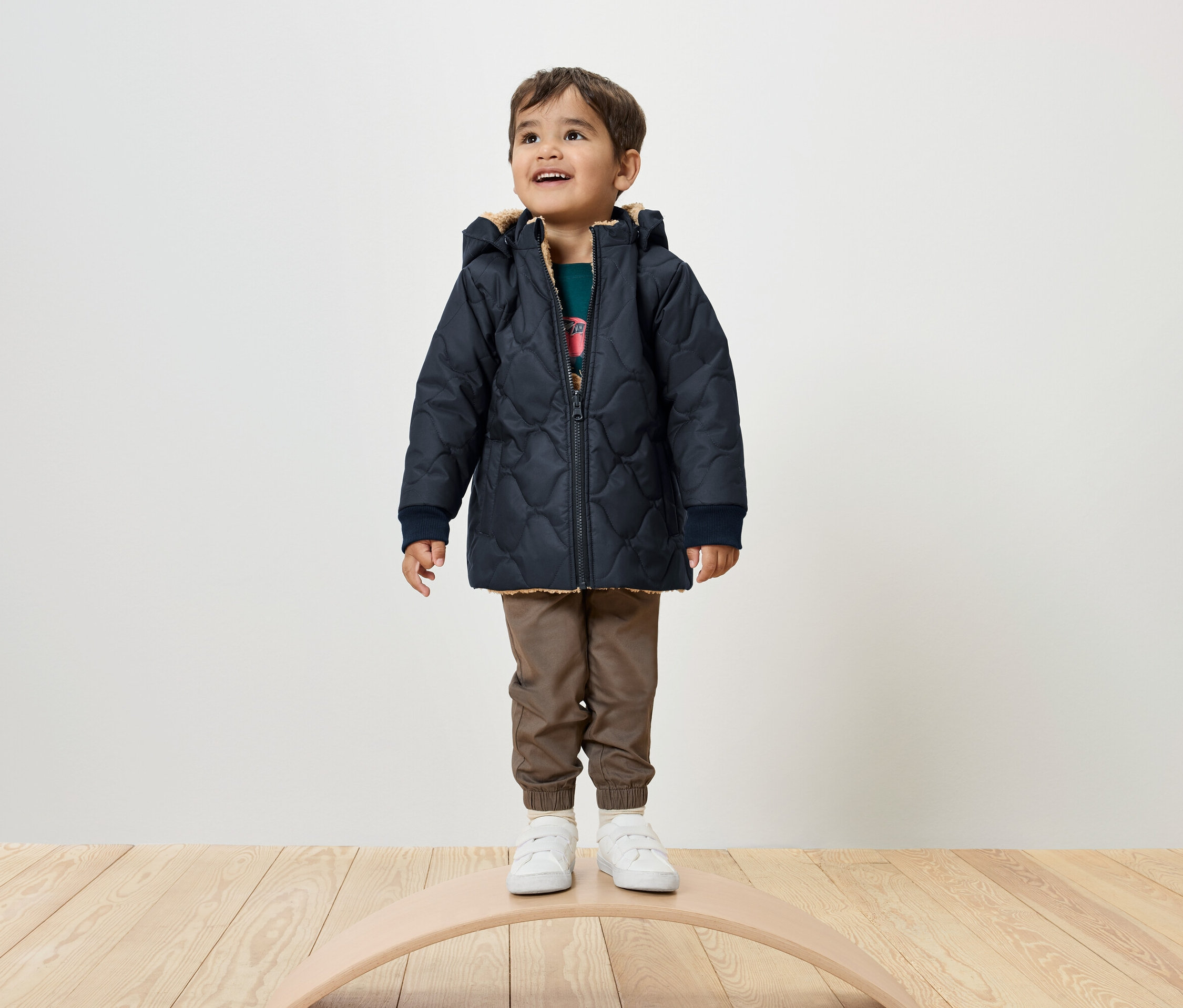 Egy fiú áll egy egyensúlyozó deszkán, sötétkék steppelt kabátot, barna nadrágot és fehér tornacipőt visel.