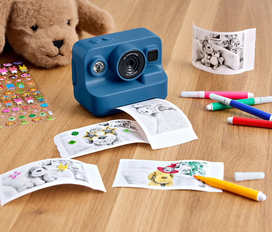 Kék nyomtatós fényképezőgép fekszik egy fa asztalon egy plüssmackóval, egy ív matricával, kinyomtatott fotókkal és színes filctollakkal.