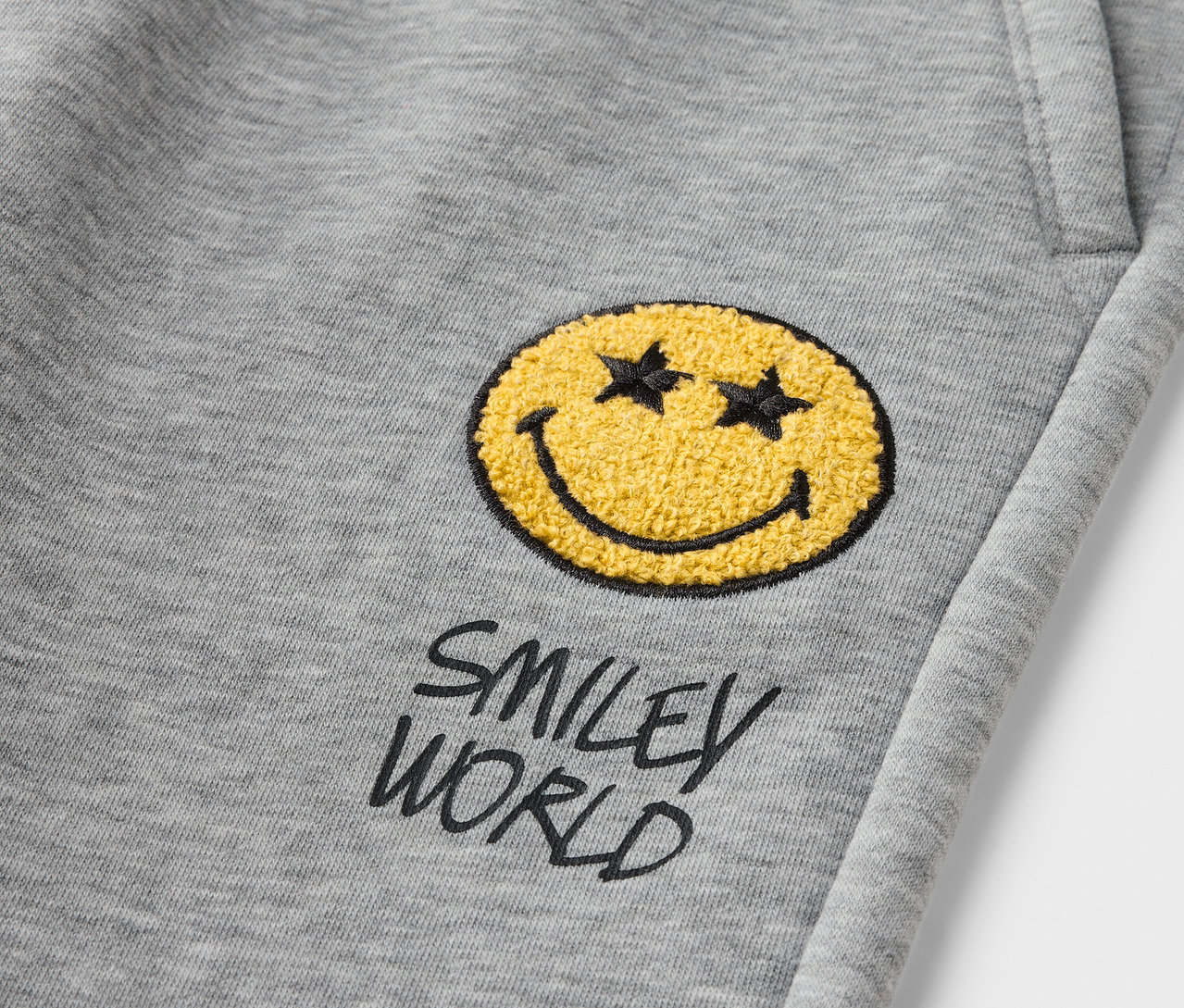 Egy szürke SMILEYWORLD® gyerek szabadidőnadrág részlete sárga mosolygó fejjel.