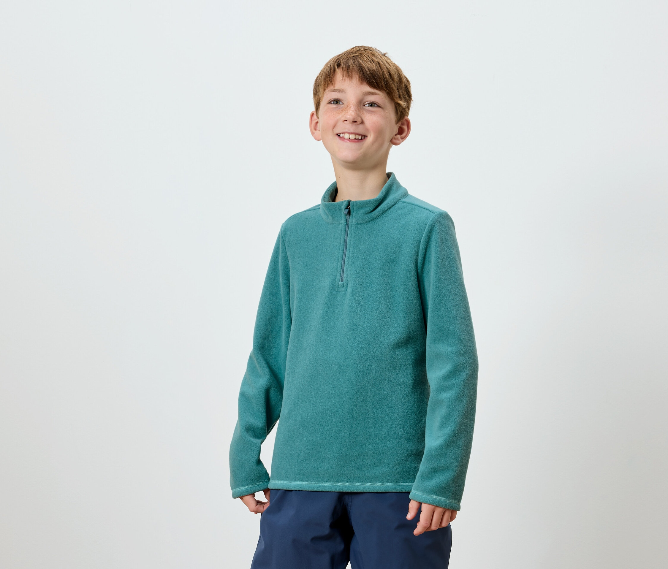 Mosolygó fiú pózol türkiz színű, cipzáras fleece pulóverben és sötétkék nadrágban.