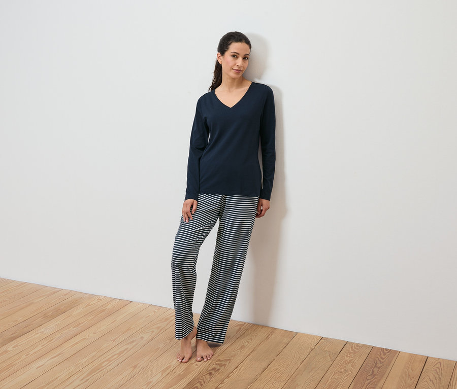 Sötétkék pizsama felsőt és csíkos nadrágot viselő nő támaszkodik egy fehér falnak.