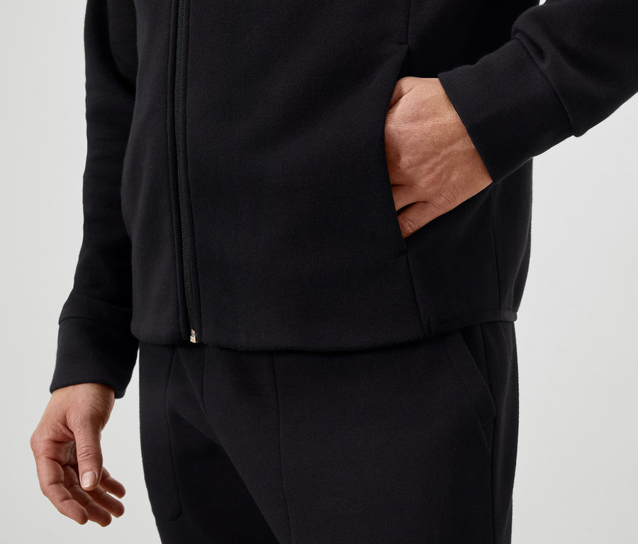 Egy férfi a kezét egy fekete, cipzáros kabát zsebében tartja, és fekete nadrágot visel.