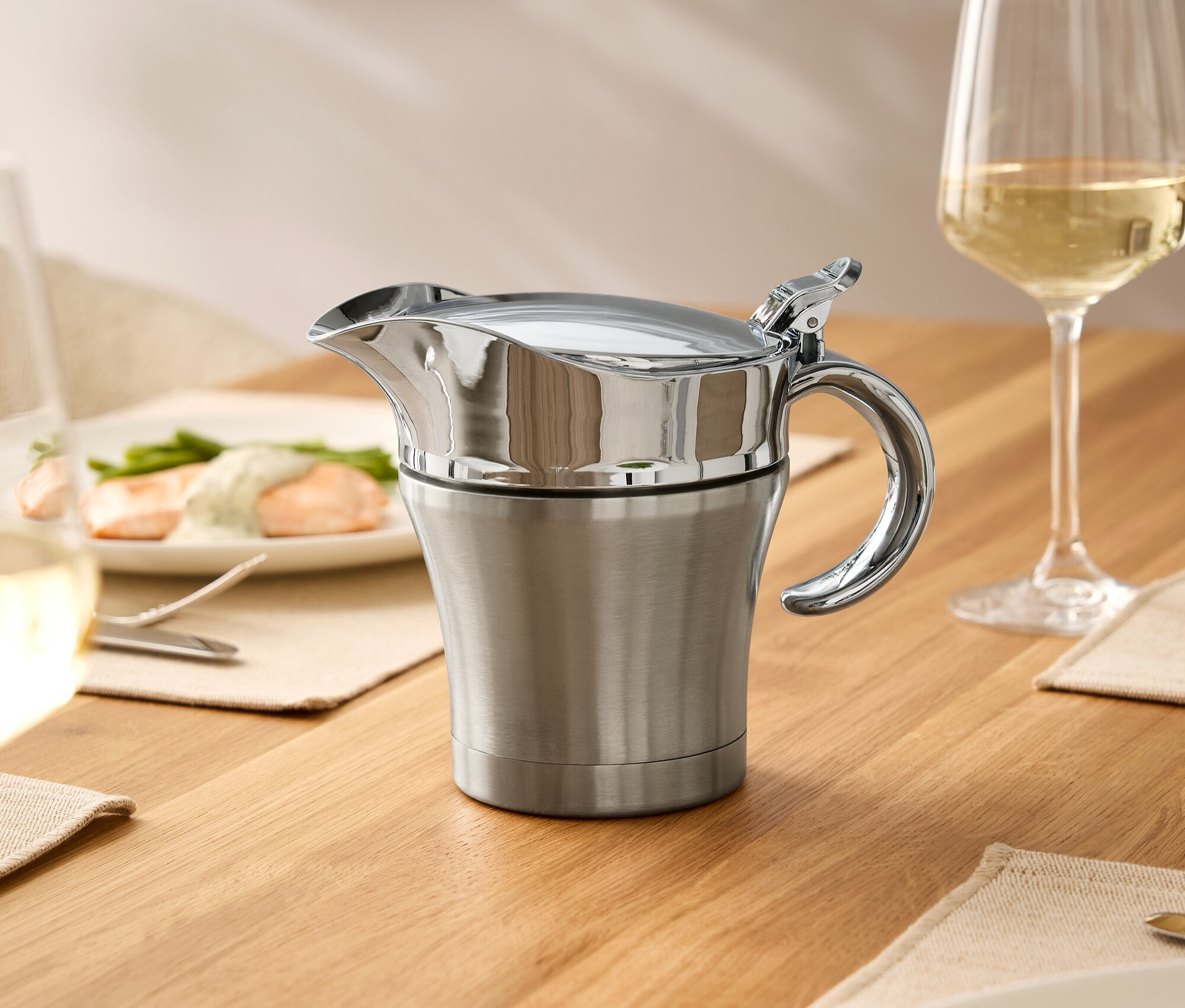 Egy fa asztalon egy rozsdamentes acélból készült fedővel ellátott mártásos edény, egy borospohár és tányérok ételekkel.