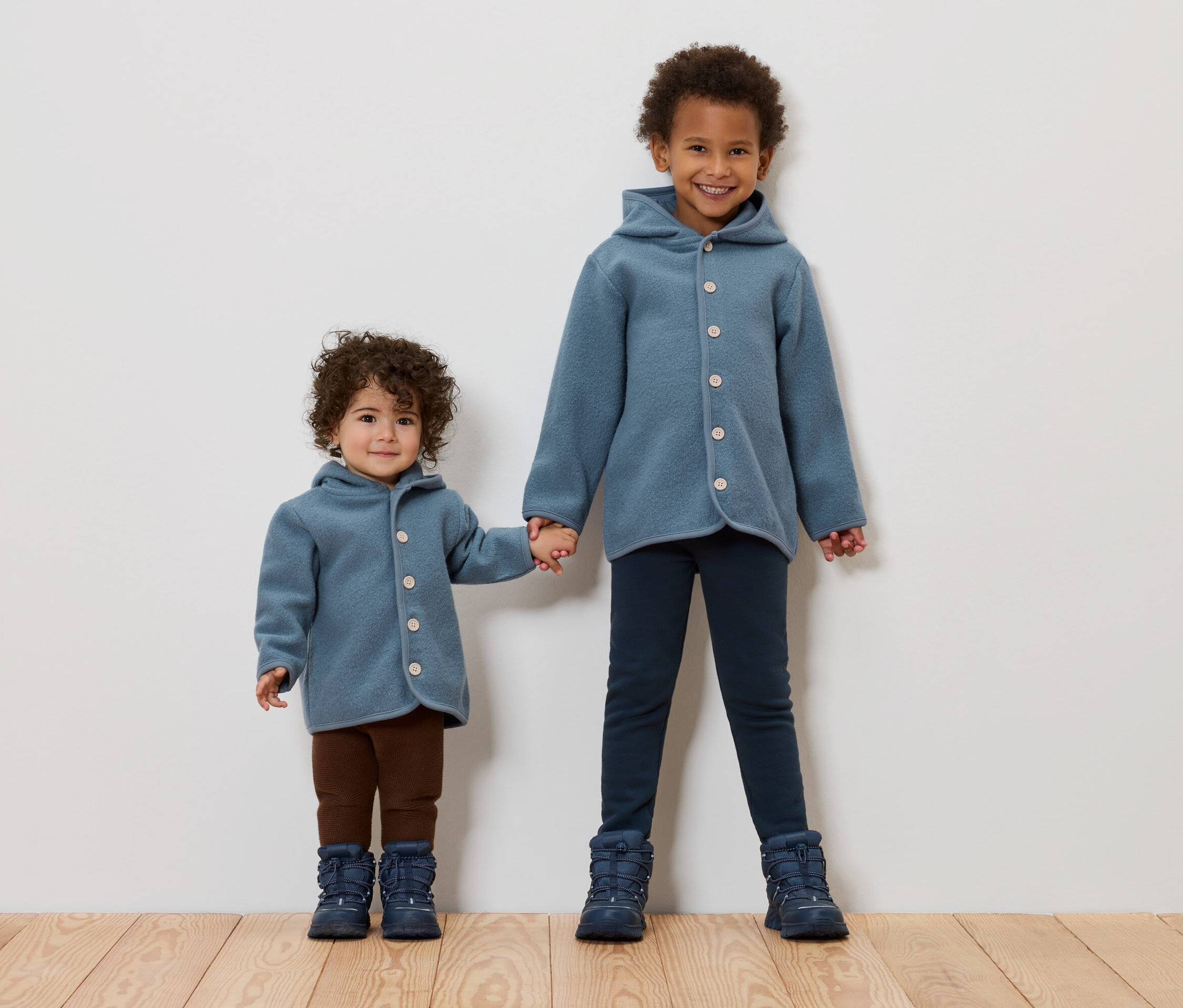 Két gyerek fogja egymás kezét. Mindketten kék kabátot és csizmát viselnek.