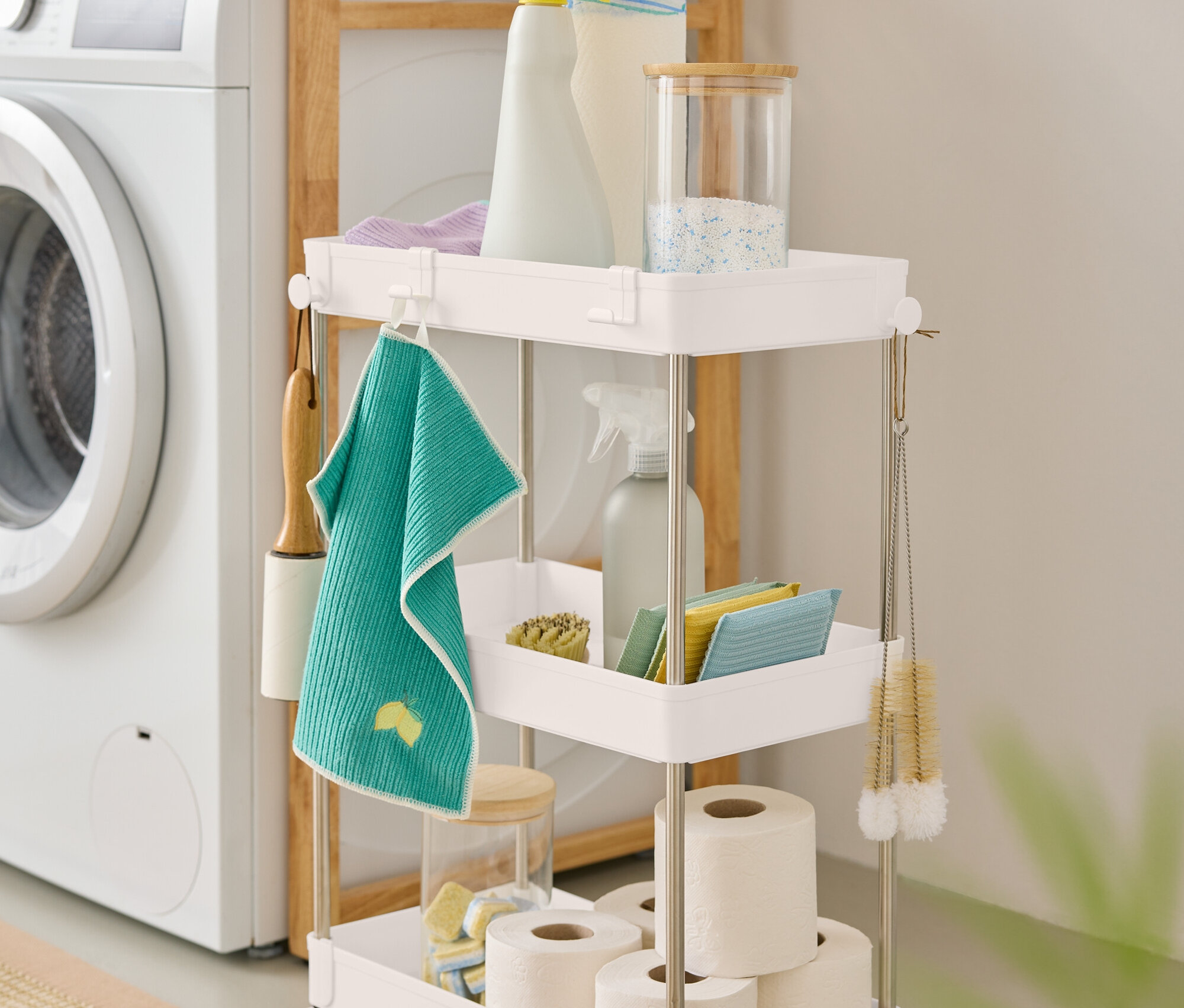 Egy háromszintes, fehér tisztítószeres kocsi áll egy mosógép mellett.