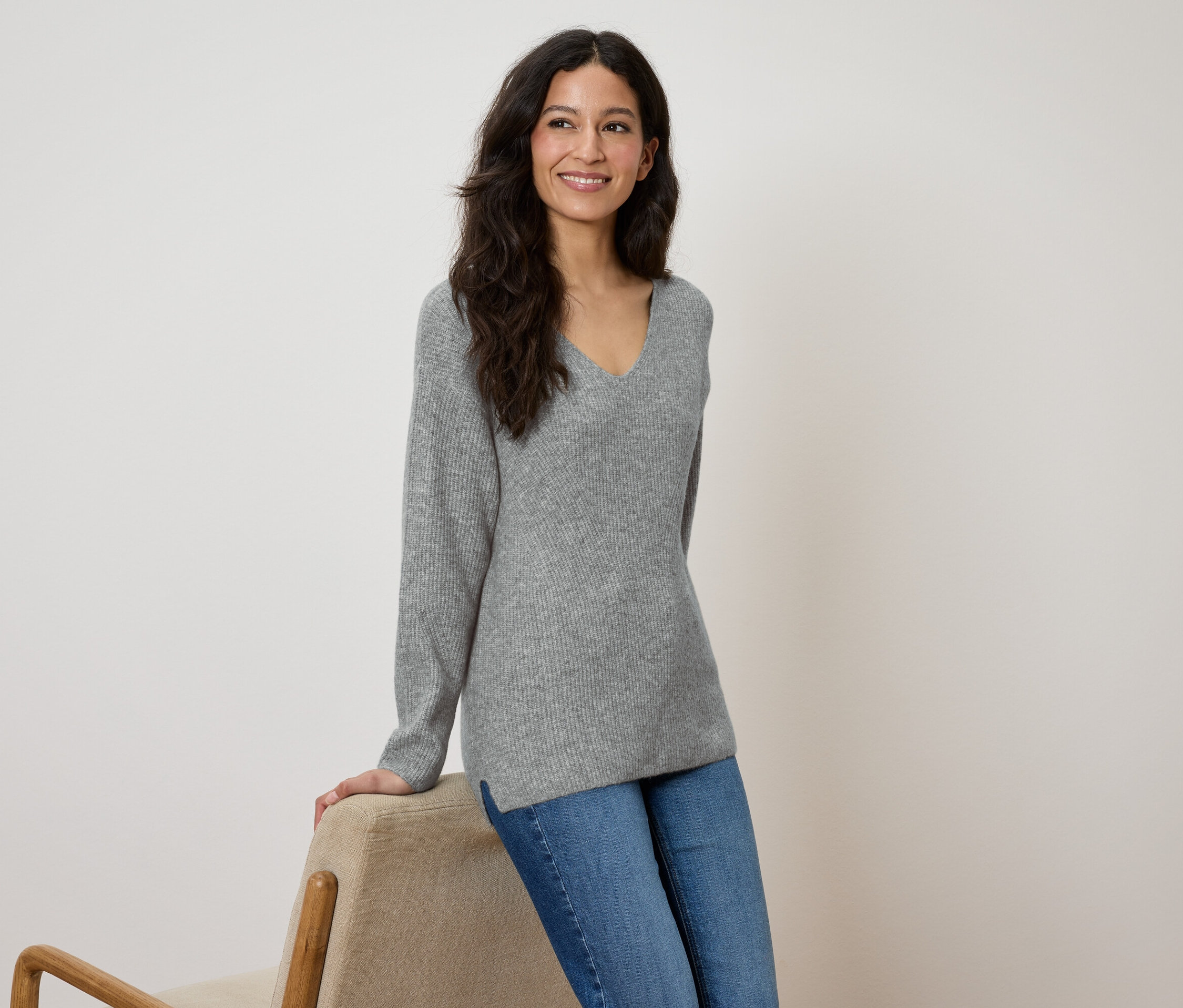 Egy nő mosolyogva támaszkodik egy bézs széknek, szürke pulóvert és kék farmert visel.