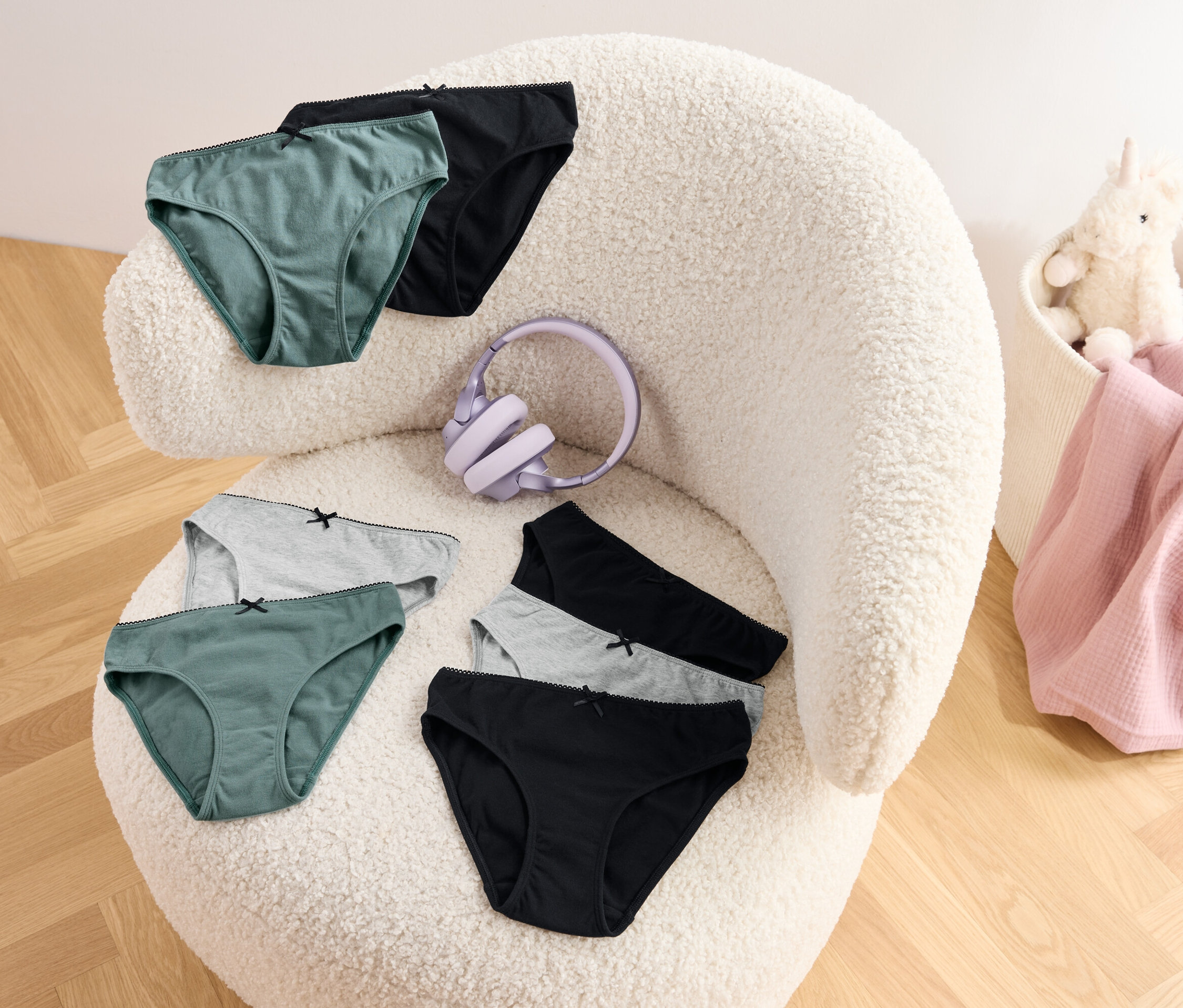 Zöld, szürke és fekete színű bugyik és egy lila fejhallgató van egy szőrös fotelre helyezve. Egy kosárban egy rózsaszín takaró és egy plüss egyszarvú található.