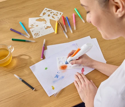 Egy nő narancssárga festéket permetez egy sablonon keresztül egy papírlapra egy airbrush toll segítségével, tollak, sablonok és egy csésze tea veszik körül.