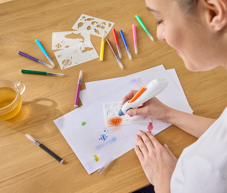 Egy nő narancssárga festéket permetez egy sablonon keresztül egy papírlapra egy airbrush toll segítségével, tollak, sablonok és egy csésze tea veszik körül.