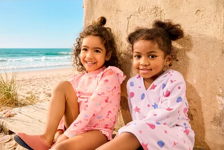 Két lány ül a strandon, az egyiken Kinder-Badeanzug mit UV-Schutzfaktor 50+ és Kinder-Badeschuhe, a másikon Leichter Kinder-Frottierponcho van.