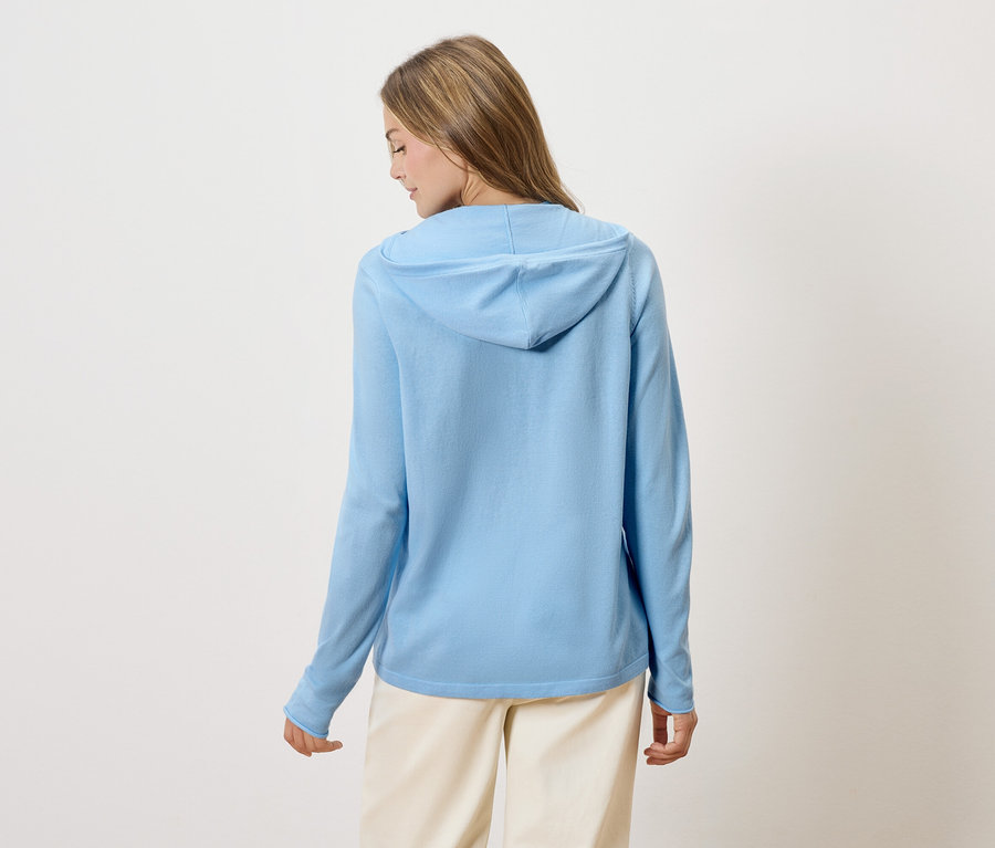 Hátulról látható egy szőke hajú nő világoskék kapucnis pulóverben és bézs színű nadrágban.