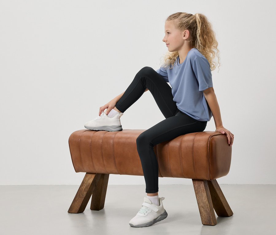 Egy szőke, lófarokba kötött hajú fiatal lány ül egy tornabakon. Kék pólót, fekete leggingset és fehér tornacipőt visel.