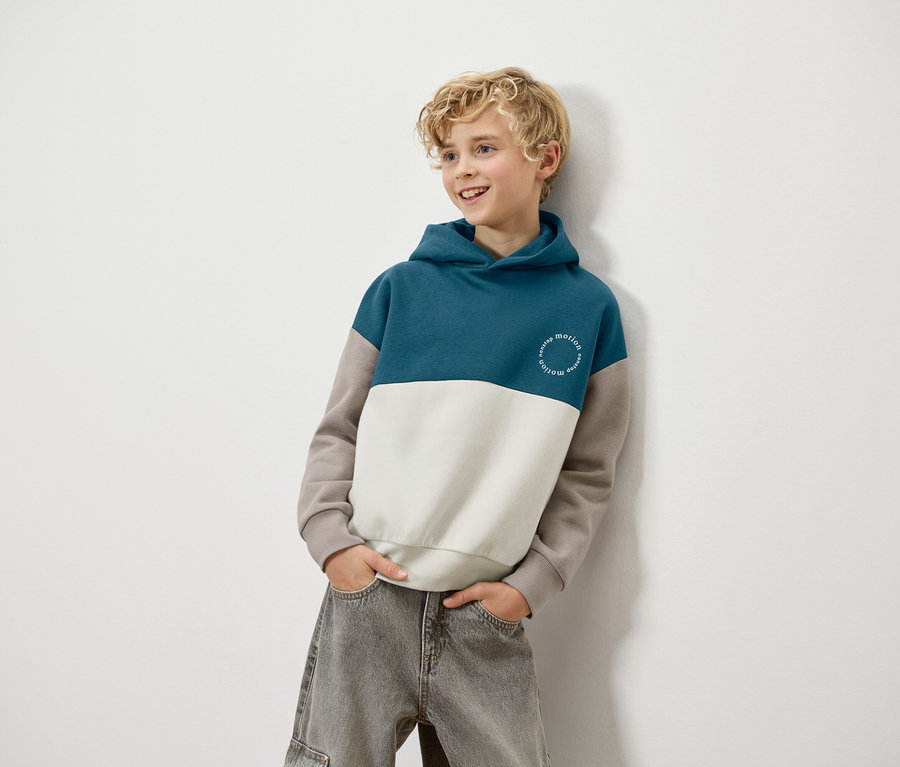 Egy szőke fiú egy falnak támaszkodik, kék, fehér és barna kapucnis pulóvert és szürke farmert visel.