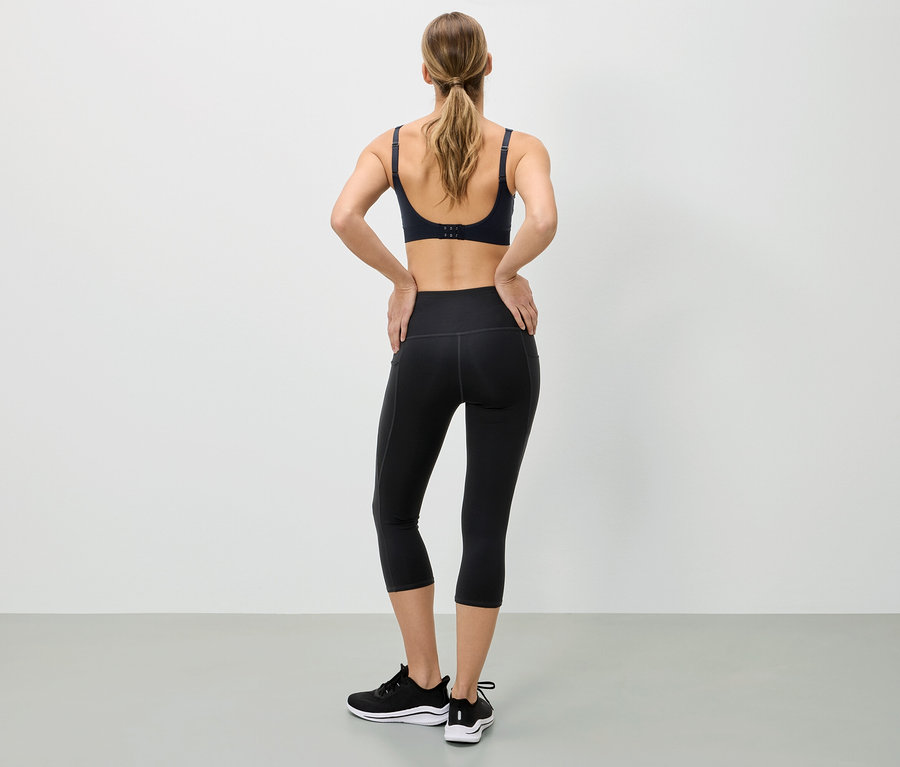 Hátulról látható nő sportmelltartóban, fekete leggingsben és sportcipőben, kezét a csípőjére helyezi.