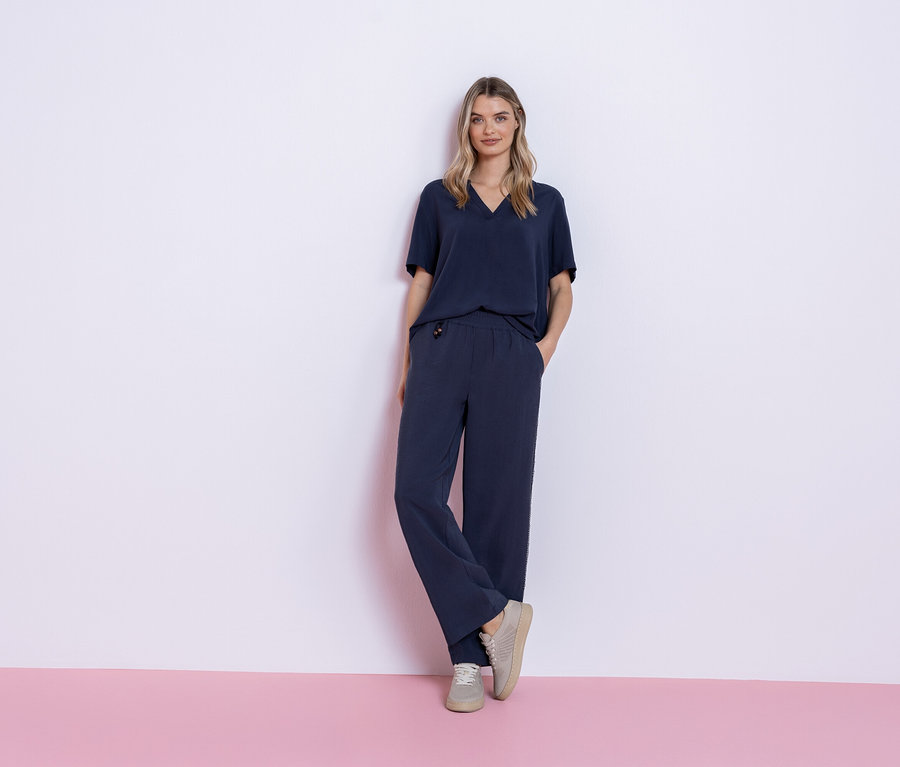 Nő áll rózsaszín és fehér háttér előtt, tengerészkék pólóból és nadrágból álló ruhát, valamint szürke tornacipőt visel.