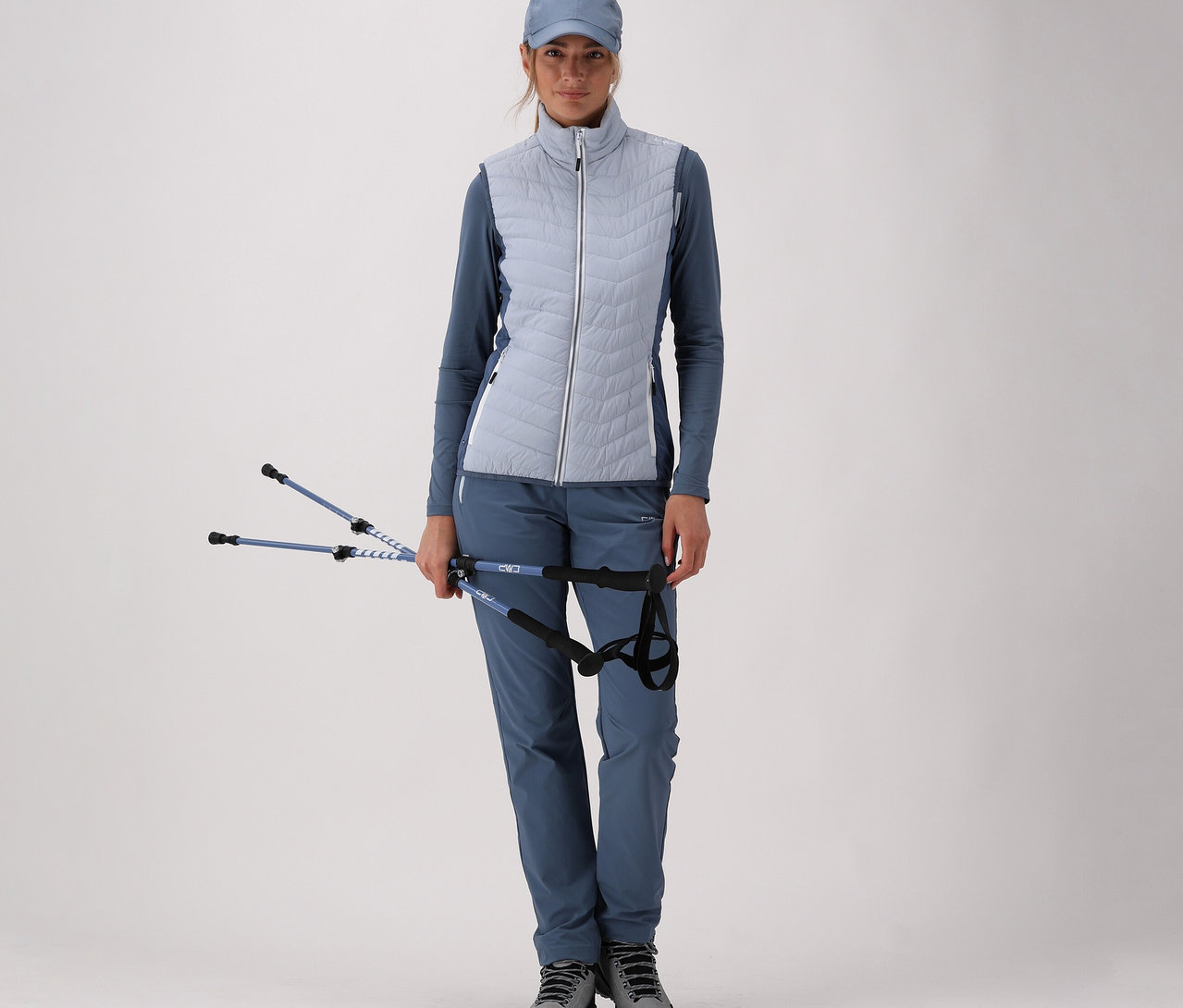 Nő kék sportruházatban, CMP női hibrid mellényben, túrabotokat tart a kezében világos háttér előtt.