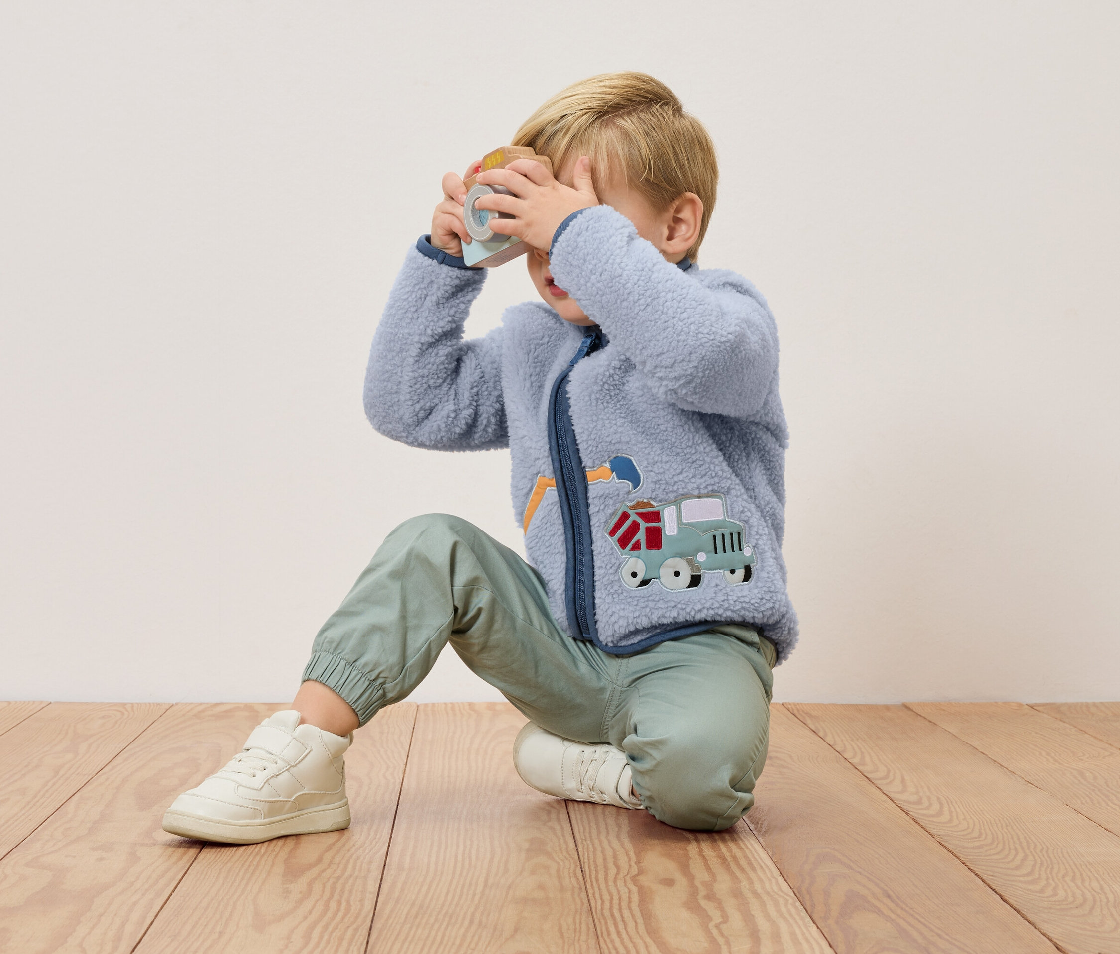 Egy szőke fiú térdel egy fapadlón, és egy játék fényképezőgépet tart az arca elé. Kék fleece kabátot visel teherautó rátéttel és zöld nadrágot.