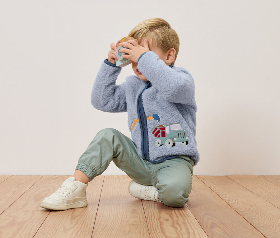 Egy szőke fiú térdel egy fapadlón, és egy játék fényképezőgépet tart az arca elé. Kék fleece kabátot visel teherautó rátéttel és zöld nadrágot.