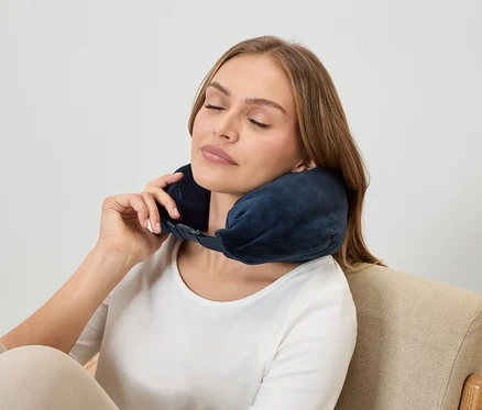 Egy nő csukott szemmel sötétkék nyaktámaszt visel, miközben egy fotelben ül.