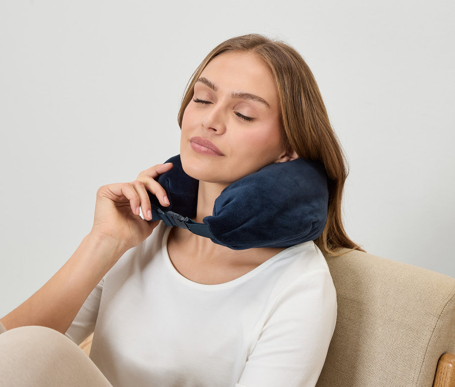 Egy nő csukott szemmel sötétkék nyaktámaszt visel, miközben egy fotelben ül.