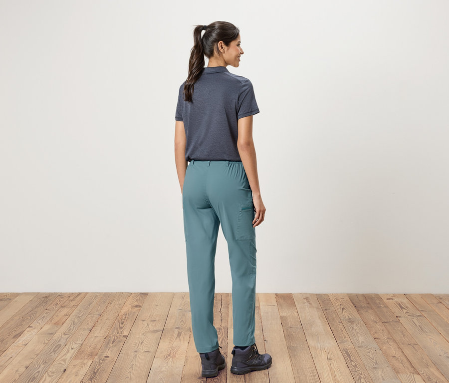 Hátulnézet egy lófarokba kötött hajú nőről, aki tengerészkék pólót, zöld nadrágot és túrabakancsot visel egy fehér fal és fapadló előtt.