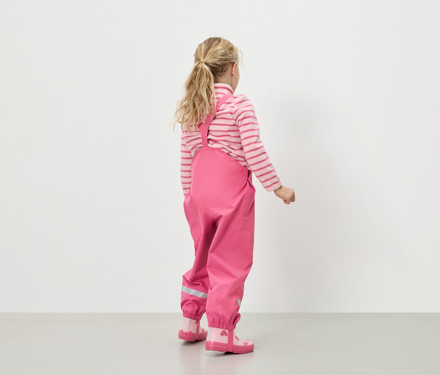Hátulnézet egy szőke hajú lányról, aki rózsaszín-fehér csíkos pulóvert, rózsaszín esőnadrágot kantárral és rózsaszín gumicsizmát visel szívekkel.