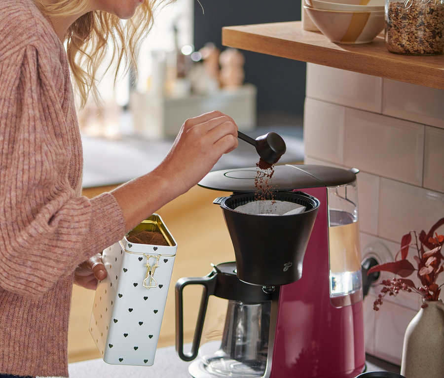 Egy szőke hajú nő kávét tölt a »Let's Brew« filteres kávéfőzőbe, berry színben.