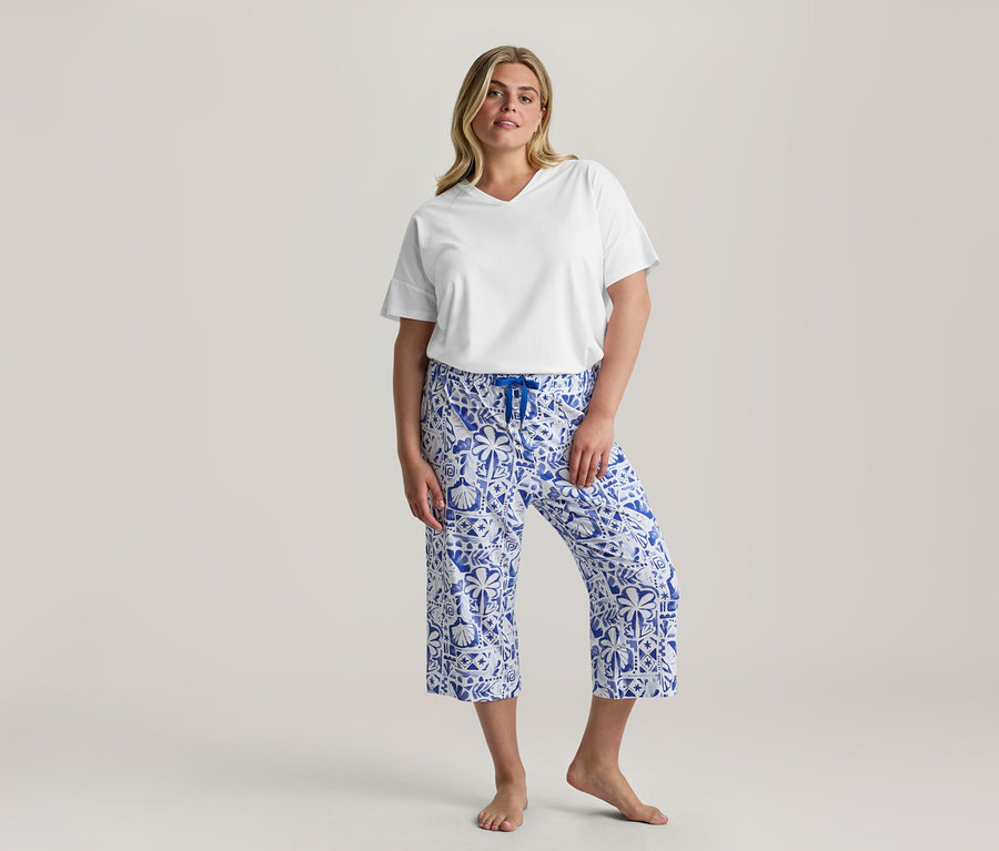 Álló nő, fehér V-nyakú pólót és kék-fehér mintás 3/4-Pyjama-Set nadrágot visel kék húzózsinórral, mezítláb.