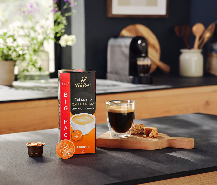 Egy csomag Cafissimo Caffe Crema kapszula, egy pohár kávé és kekszek egy asztalon. A háttérben egy Cafissimo pure+ black kávéfőző.