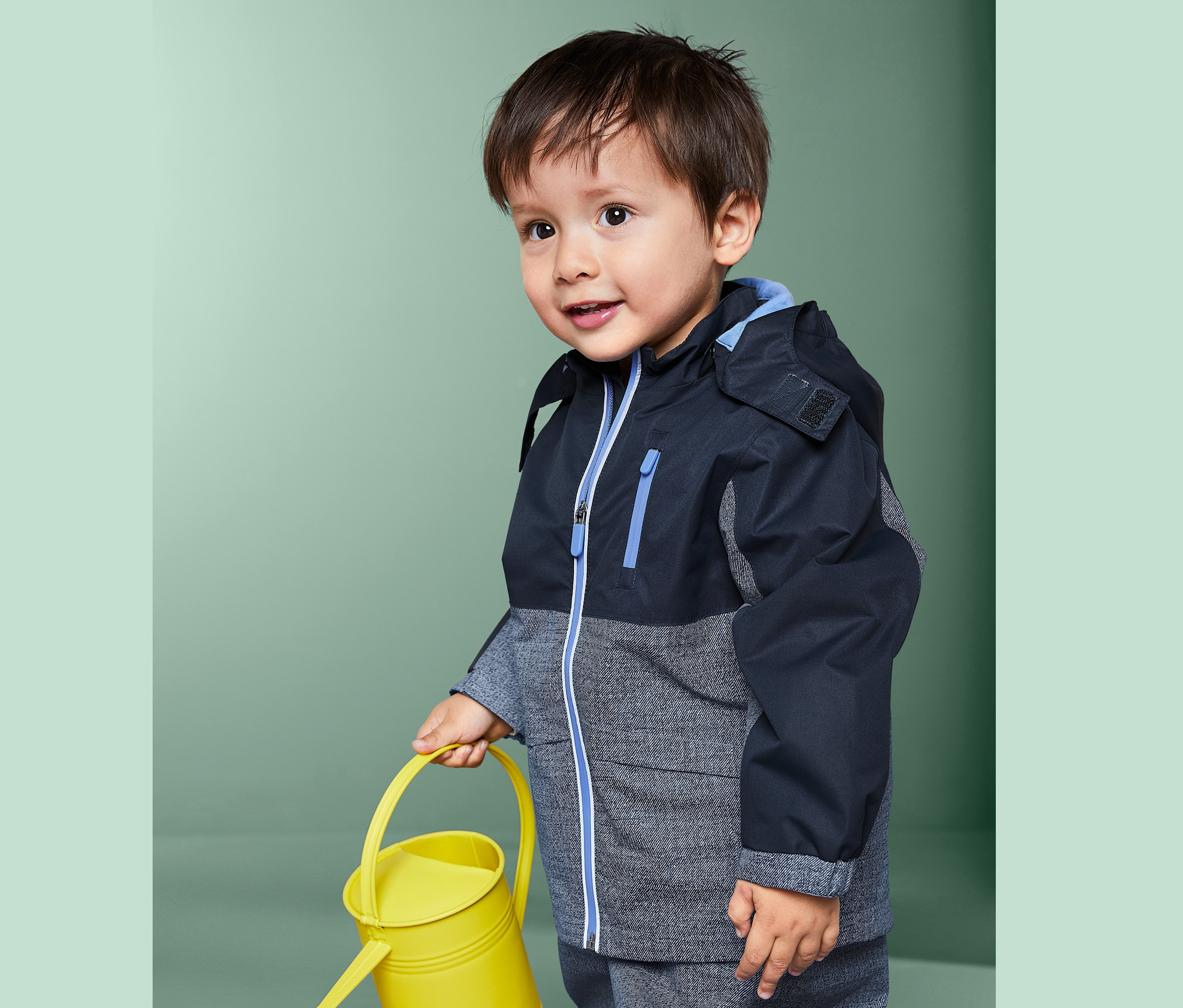 Kisfiú sárga locsolókannát tart és Gyerek bélelt esőkabátot visel.