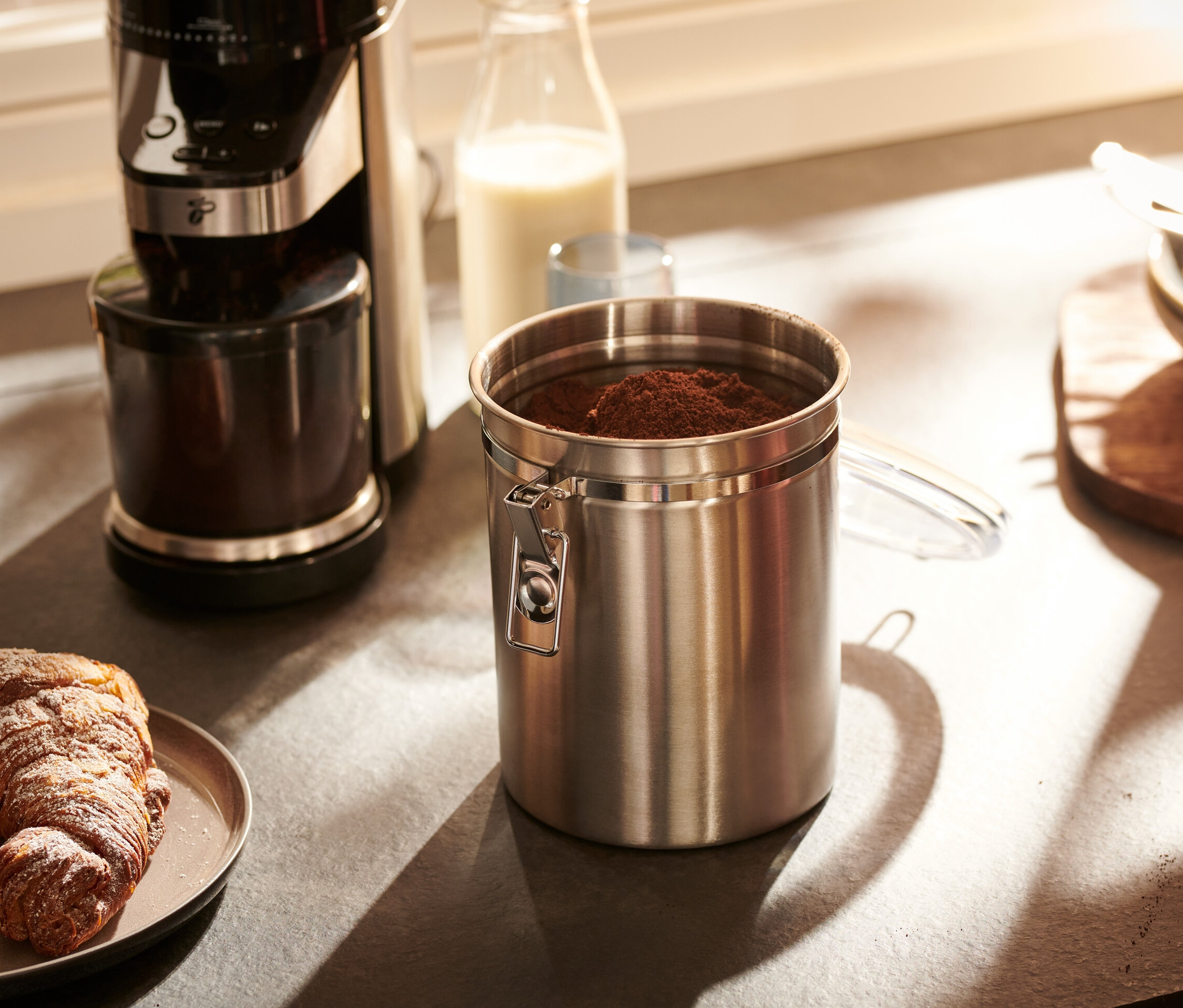 Egy rozsdamentes acélból készült aromatartó doboz áll a konyhapulton egy kávéfőző és egy croissant-t tartalmazó tányér mellett.