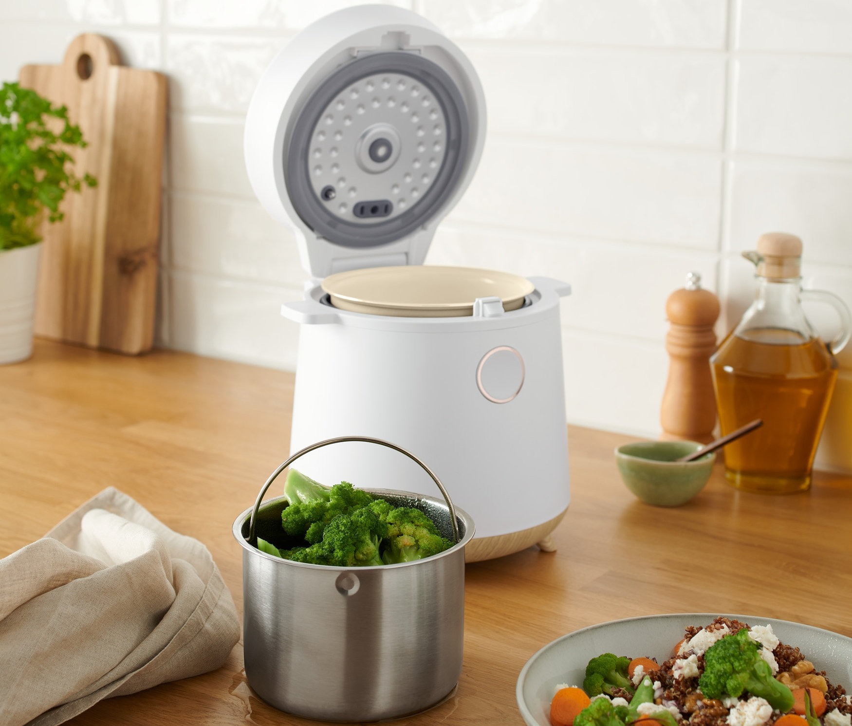 Egy fa felületen egy nyitott multifunkcionális főzőedény áll, mellette egy tál brokkolival és répával.