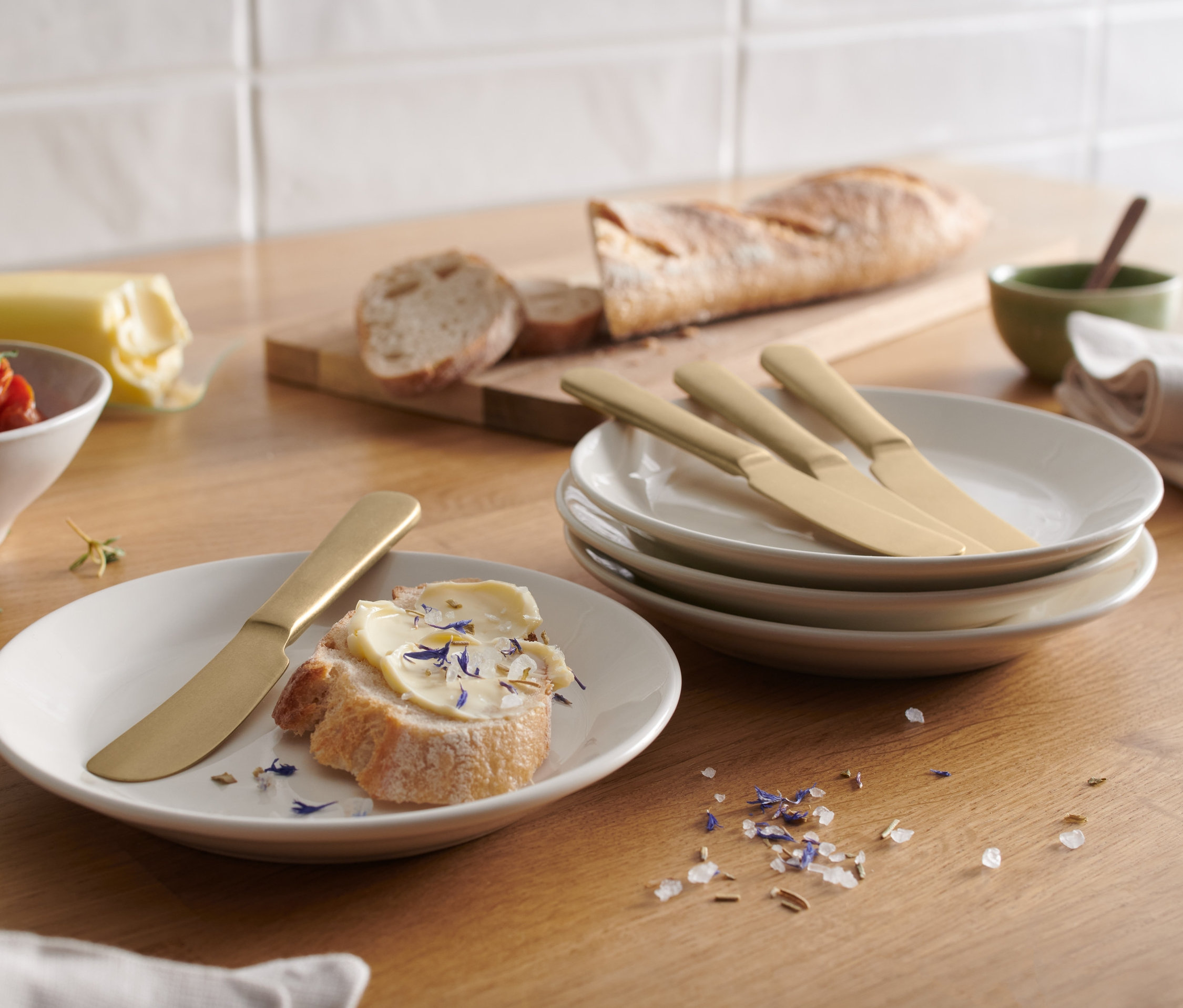 Az asztalon kenyér vajjal és egy vajazókés, mellette 4 vajazókés tányérokon.