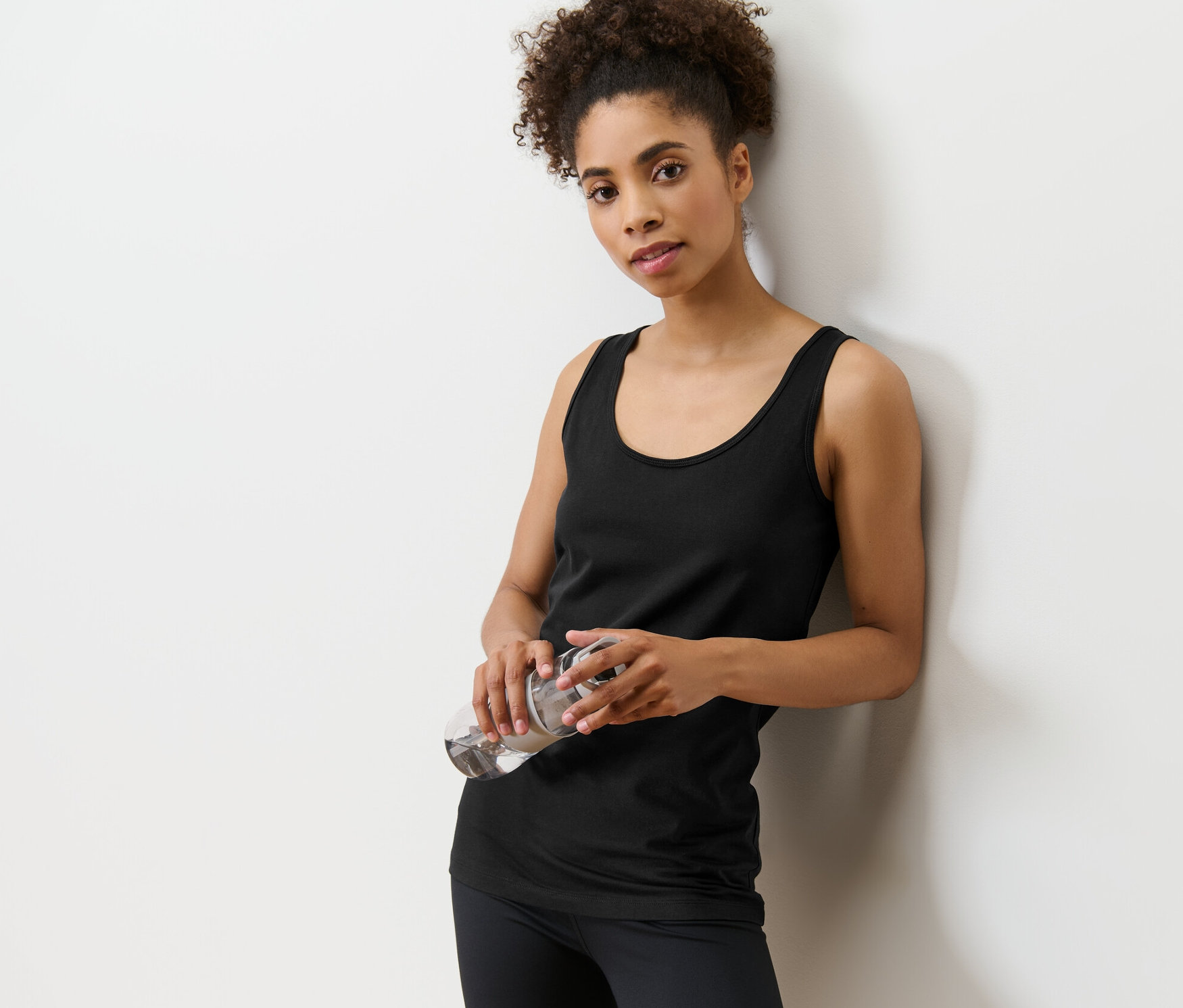 Falnak dőlve vizet tartó nő. Fekete topot visel egy kétdarabos szettből.