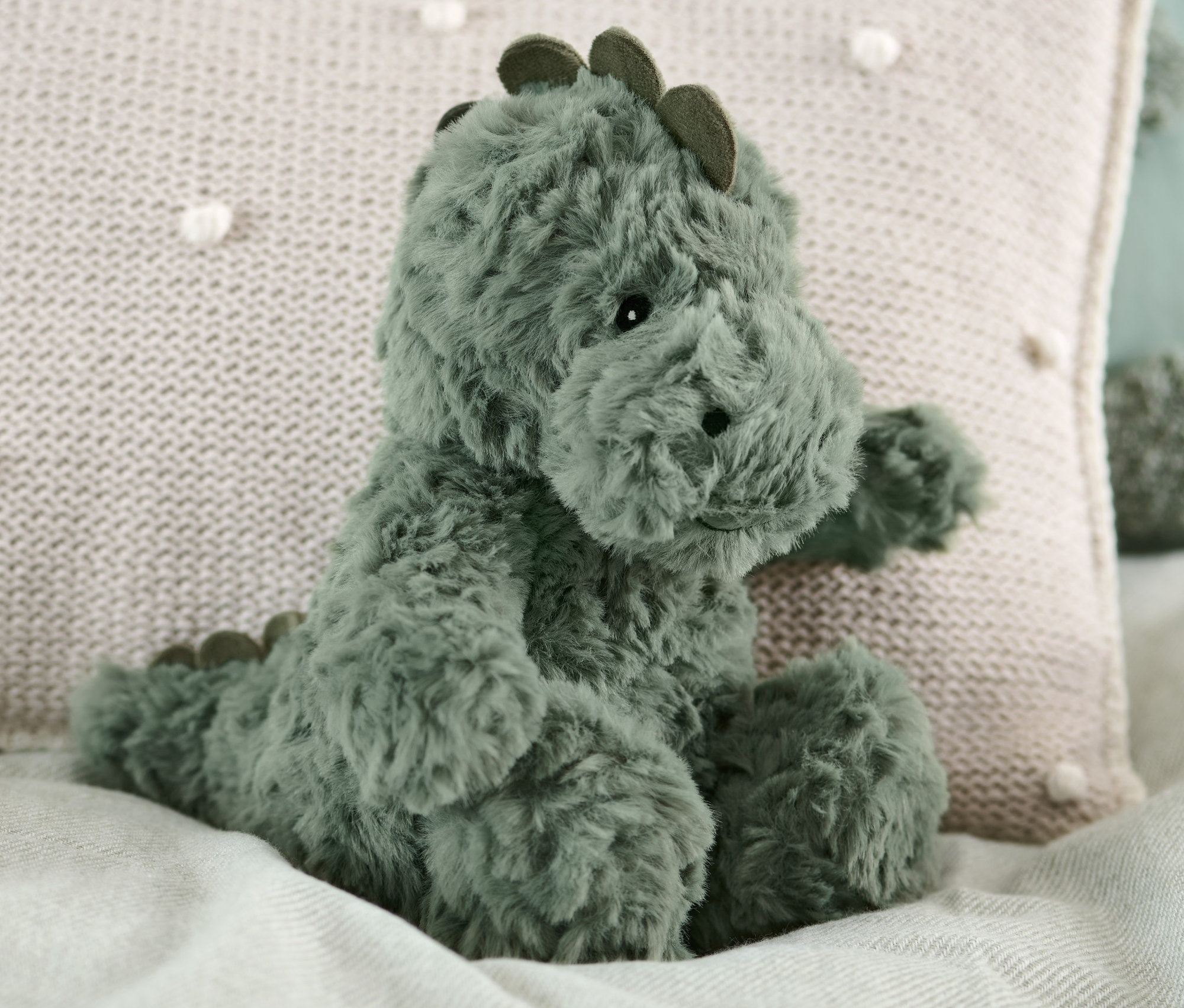 Egy plüss dínó ül egy ágyon egy bézs párna mellett.