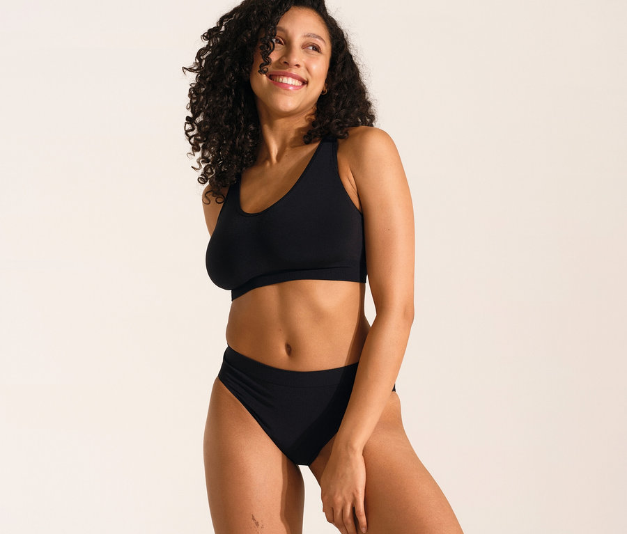 Mosolygó nő pózol fekete, betétes SCHIESSER puha melltartóban és fekete SCHIESSER Rio fazonú alsóban.