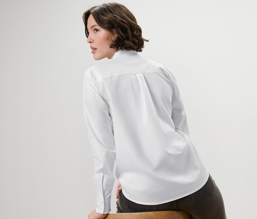 Nő ül oldalra egy fa széken, fehér, hosszú ujjú poplin blúzt visel.