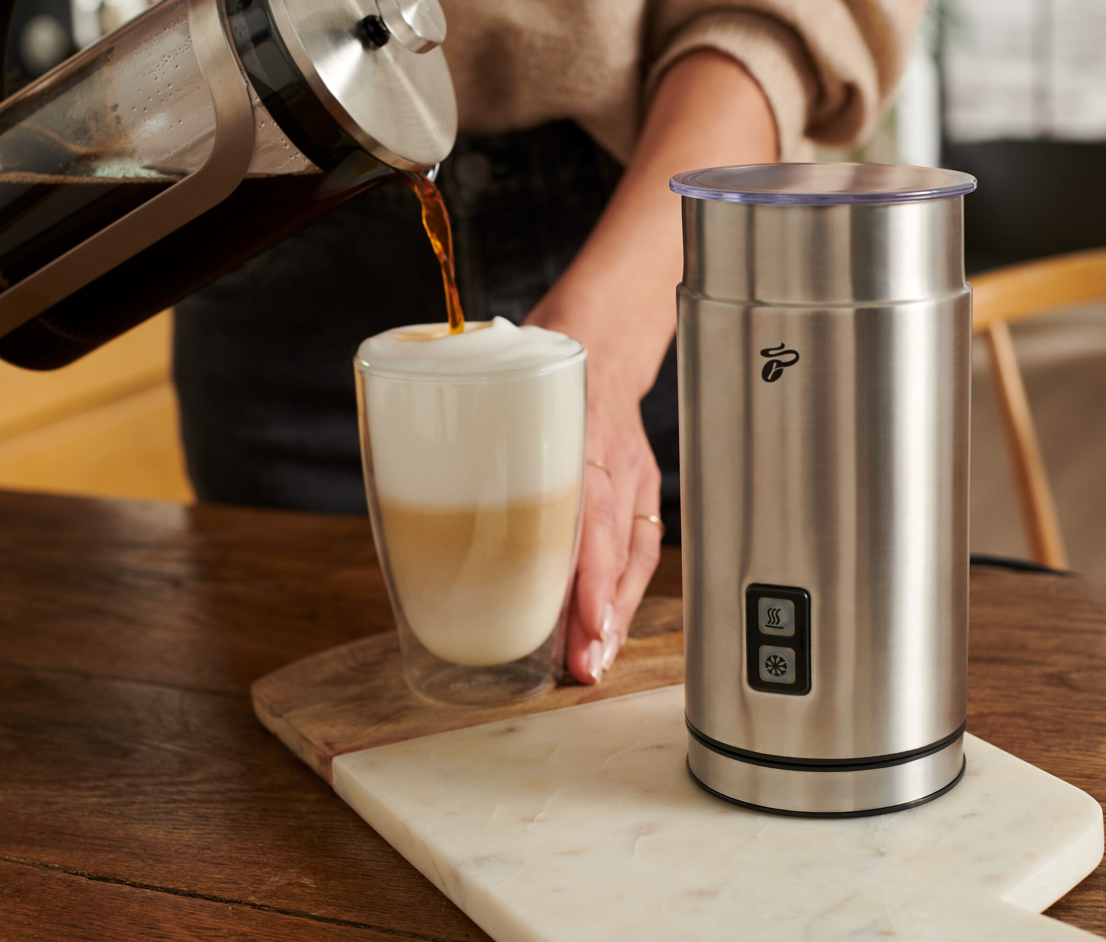 Nő kávét önt egy tejes pohárba egy elektromos tejhabosító, rozsdamentes acél mellett.