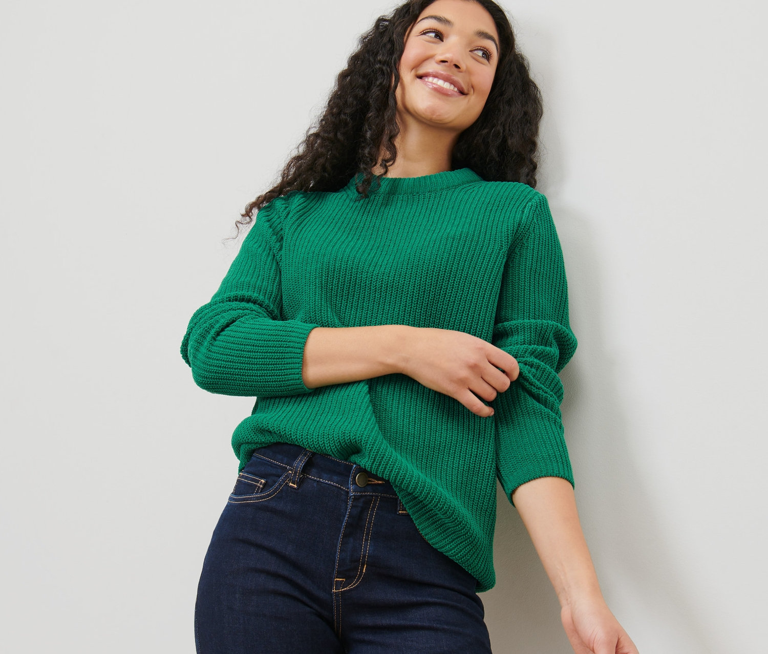 Sötét, göndör hajú nő visel egy zöld, laza kötésű pulóvert és farmert.