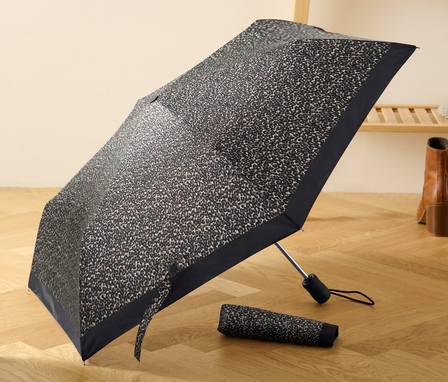Egy nyitott, leopárdmintás automata táskaesernyő fekszik egy fa padlón, egy összehajtott esernyő mellett.